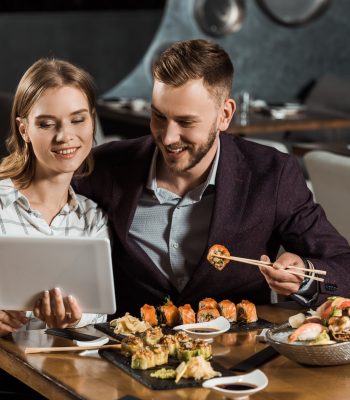 Attractive couple of yound adult using digital tablet while eating sushi in restaurant